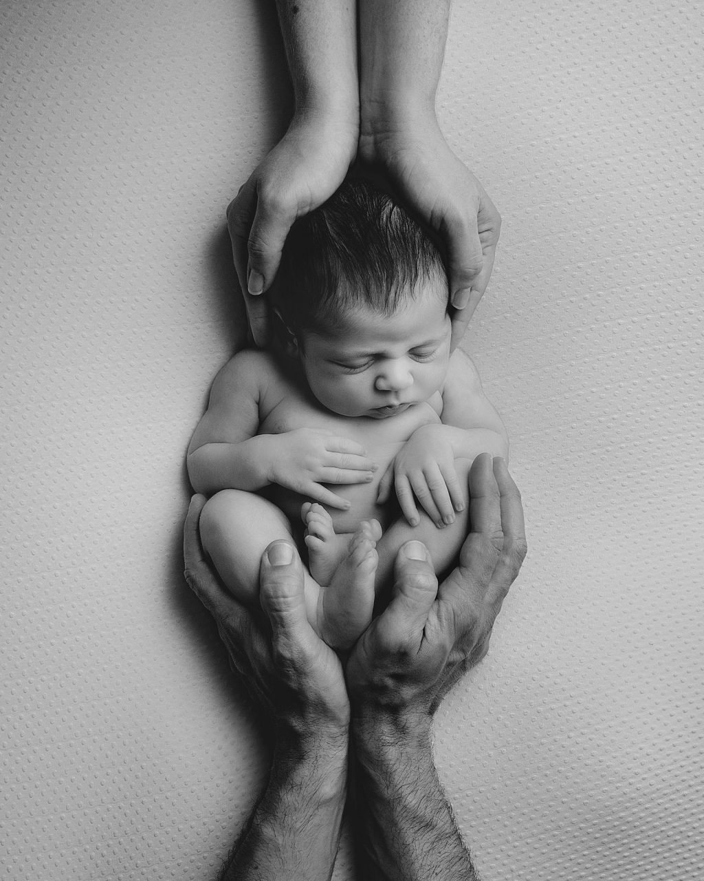 black and white image of a newborn baby on soft cushion with a pair of male hand cupping them from below and female hands cupping them from above