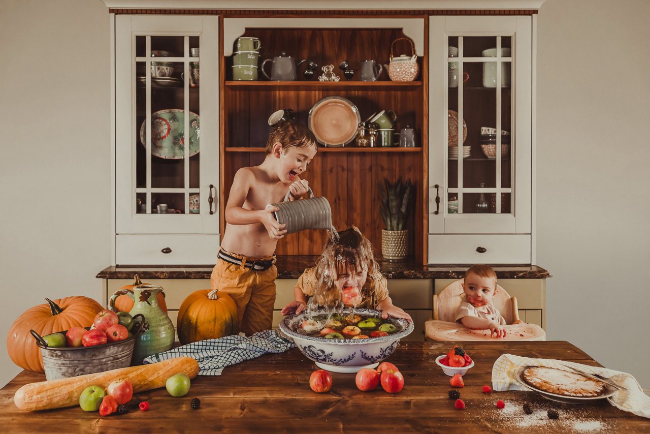 Three children in the kitchen, at a countertop. One child is dunking for apples in a large bowl, while a slightly older child is pouring a jug of water over them. The third child, a toddler in a high-chair, looks over their shoulder at their siblings.
