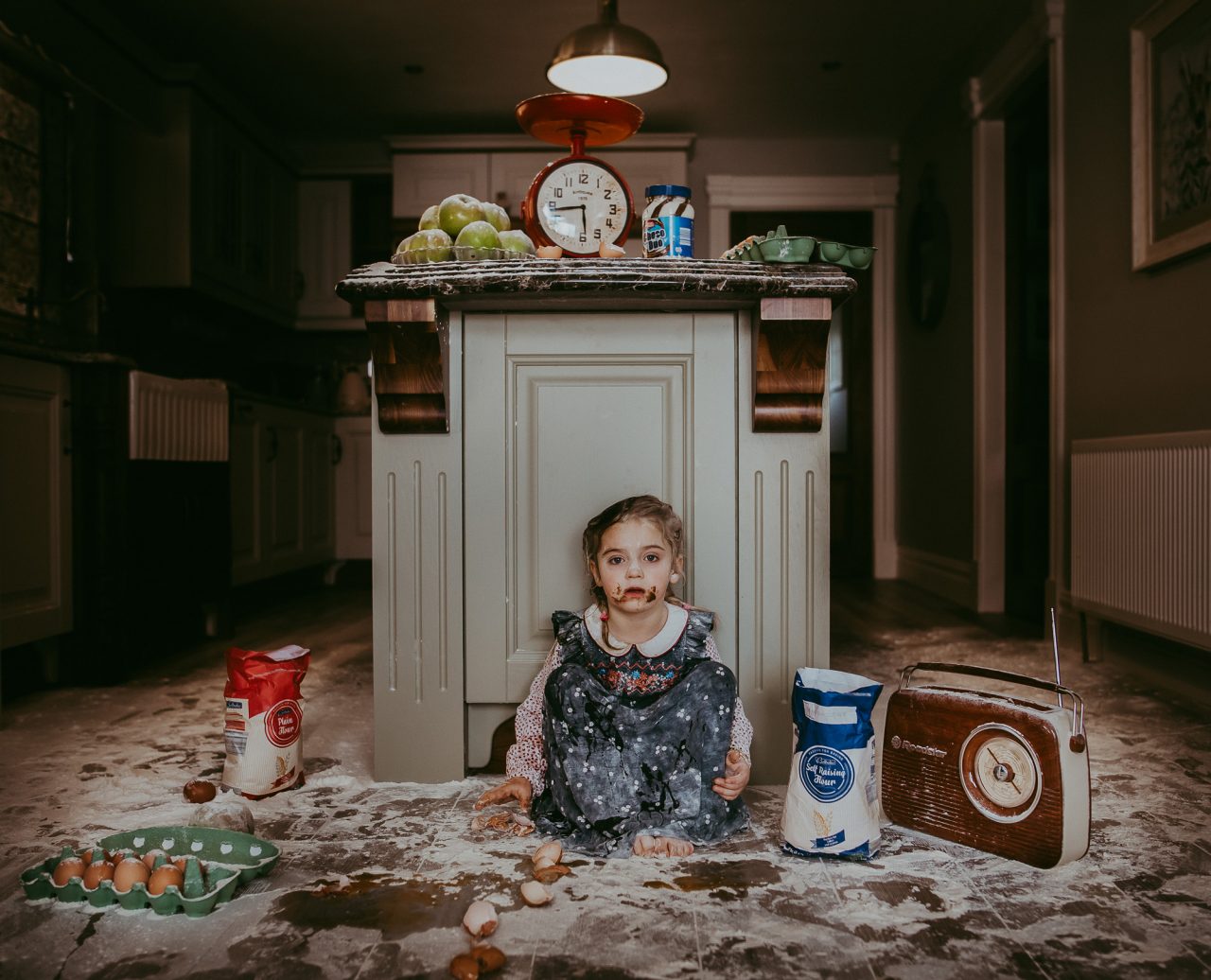 Kitchen scene of a little girl sat on the floor, staring into the camera, surrounded by flour and other baking goods