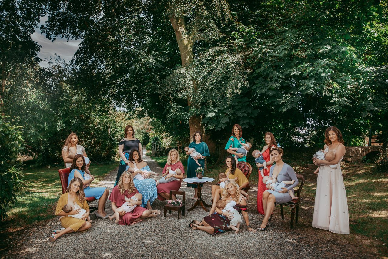 Fourteen women are posed on a pebble garden path surrounded by large trees, on a bright and sunny day. They are all holding and breastfeeding their babies, and they are all dressed in fashionable evening gowns 