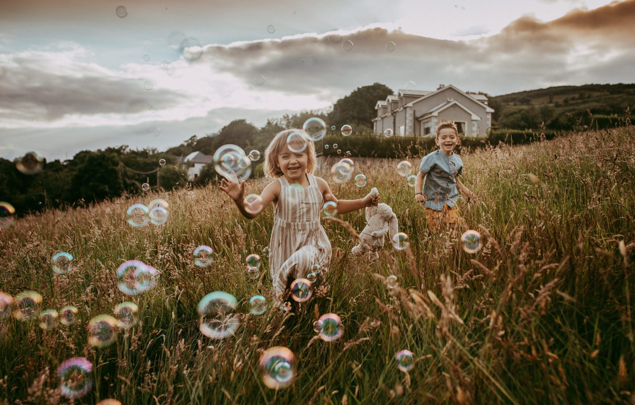 two children running through a field of tall grass, chasing bubbles