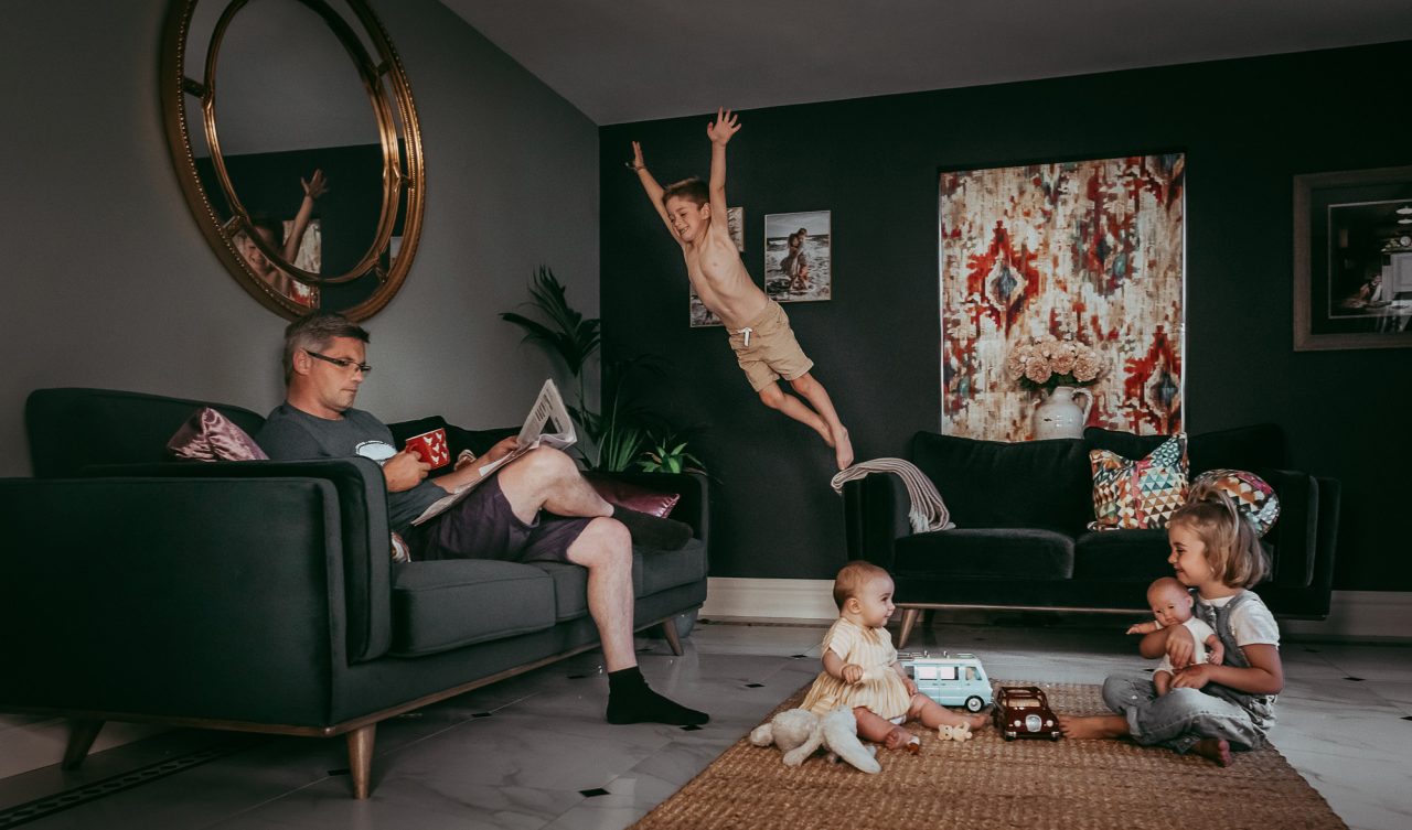 living room family scene. The father is relaxing with a newspaper and a cup. One child is mid-air, jumping from one couch to the couch where his father is seated. The other two children are playing on the ground