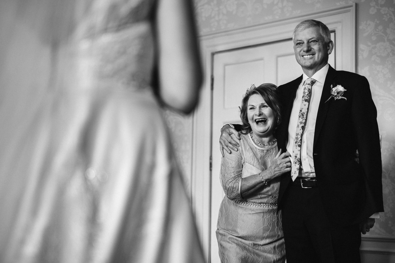 a bride, with her back to the camera, faces her joyful parents, who are embracing while they look at her