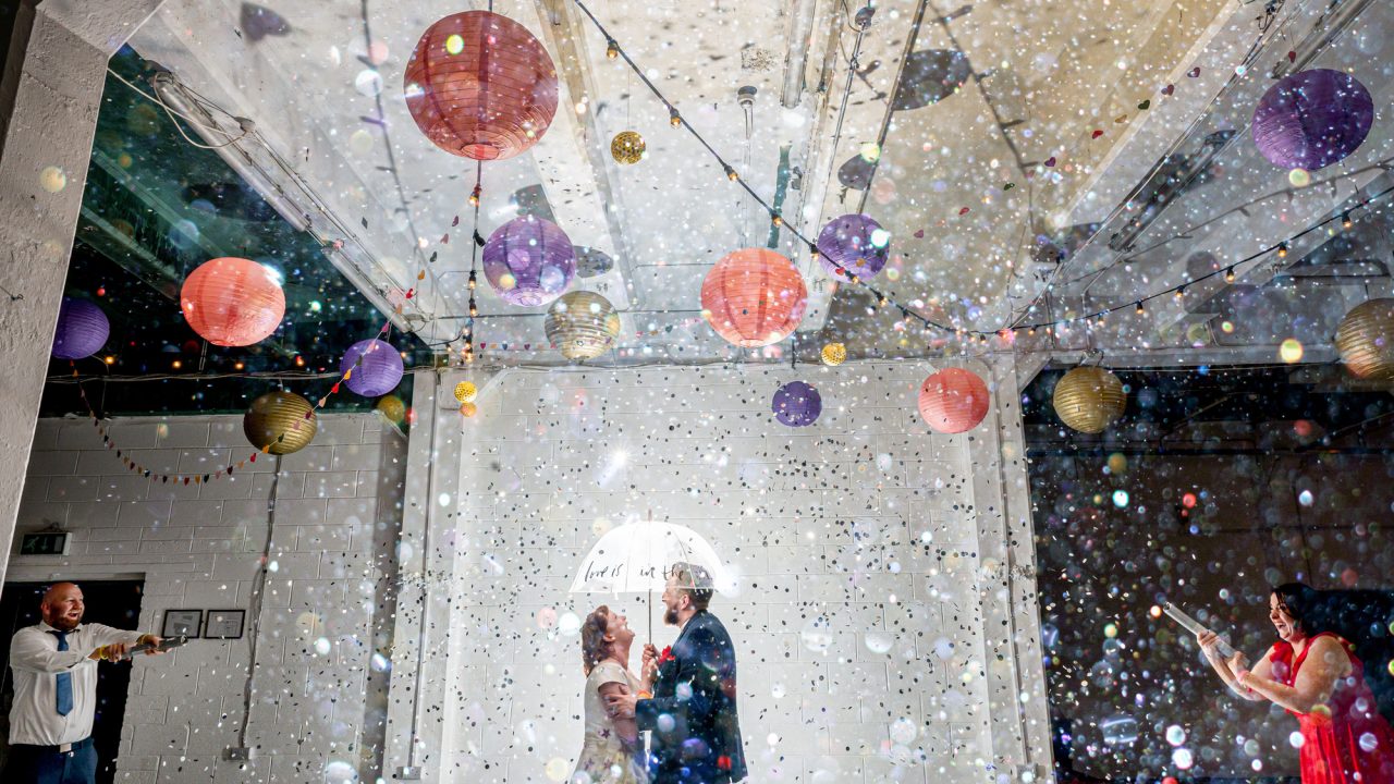 bride and groom under a small white umbrella in a large room exploding with confetti and paper lanterns hanging from the ceiling. Two wedding guests are at either side with confetti canons