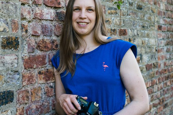 female photographer with blue top leaning against a brick wall with a camera in her hands