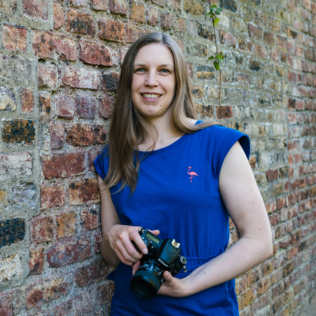 female photographer with blue top leaning against a brick wall with a camera in her hands