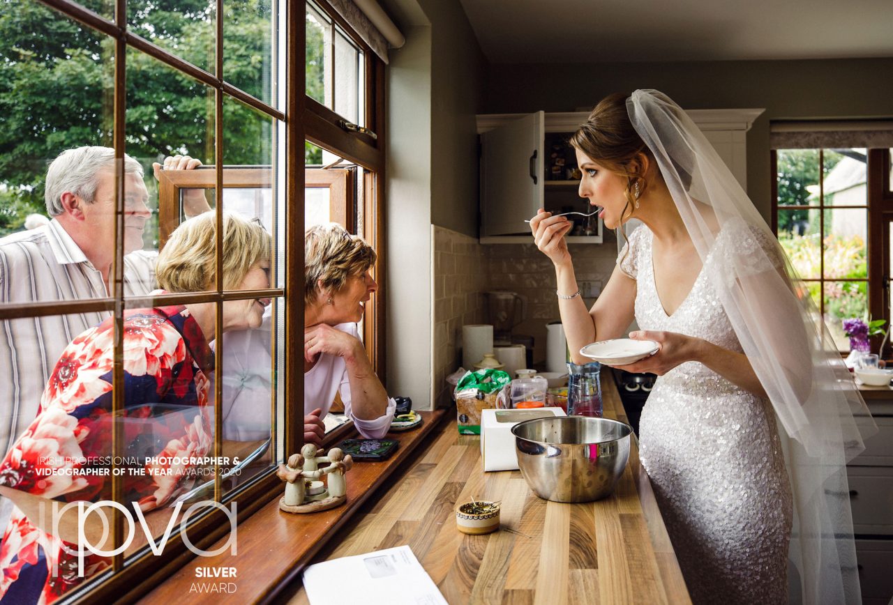 award-winning image of a bride to be eating some cake inside a kitchen while talking to older relatives who are sticking thier heads through a window from outside